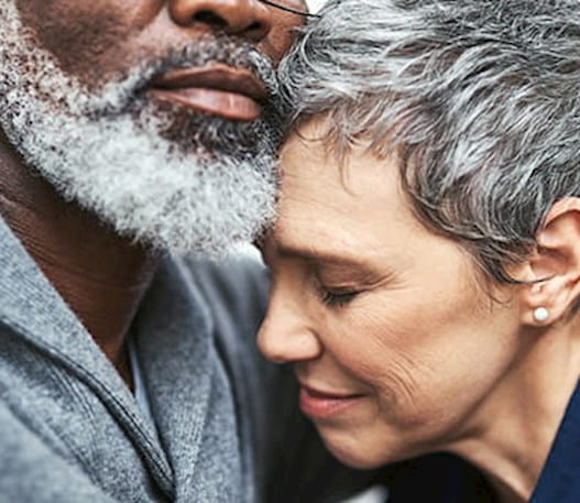The face of a woman with short grey hair looking sad, hugging a man with a beard