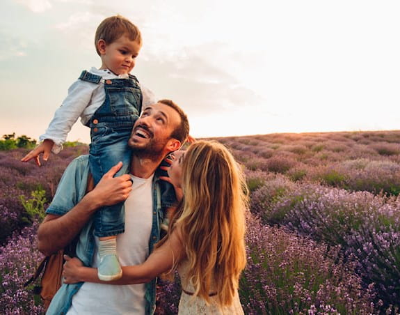 A man with a beard holding a small child in overalls which a woman with long hair embraces him. Lavender field in the background.
