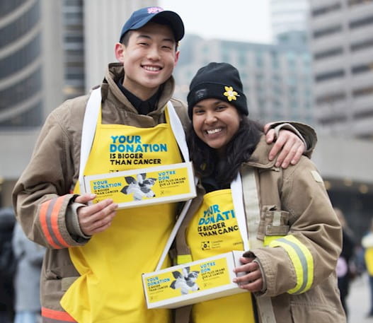 Young man and young woman wearing yellow Daffodil campaign aprons,  holding daffodil pin boxes at City Hall, Toronto
