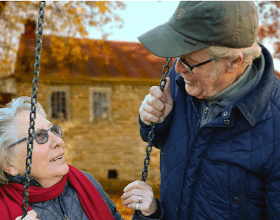 An elderly man and woman are smiling at each other while the woman sits on a swing