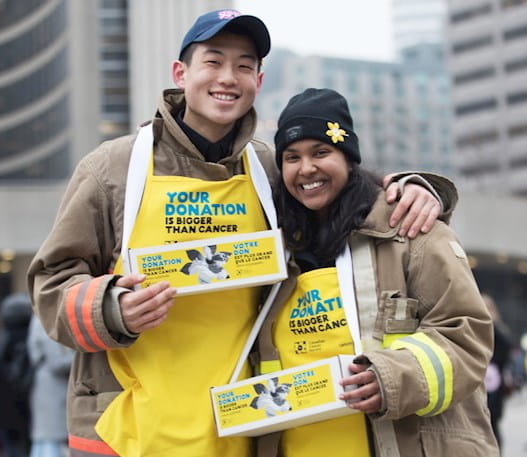 Jeune homme et jeune femme portant des tabliers jaunes de la campagne de la jonquille et tenant des boîtes d'épingles à jonquille à l'hôtel de ville de Toronto.